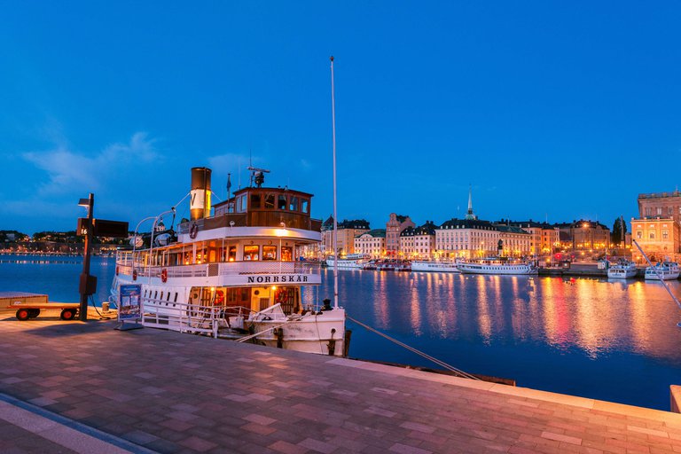 An old steamer boat lies moored in the waters outside of Stockholm Palace. Evening. The Old Town is visible in the background.