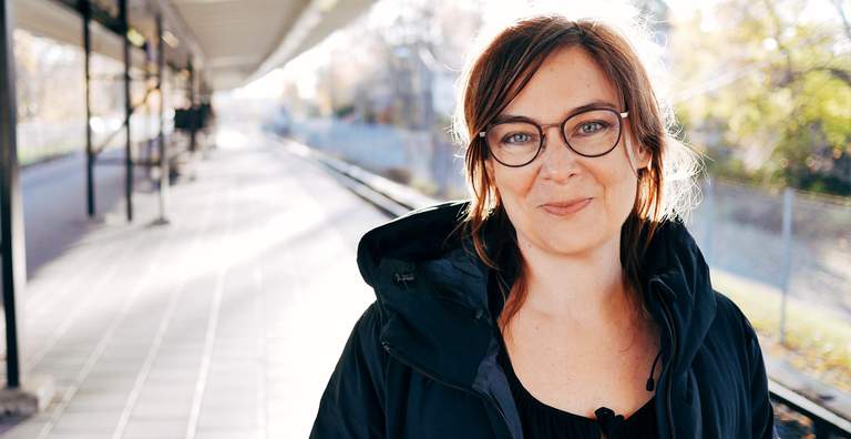 Spring in Stockholm. A middle-aged woman, standing outside on a subway platform, is smiling into the camera. The woman is Maria Miesenberger, artist.