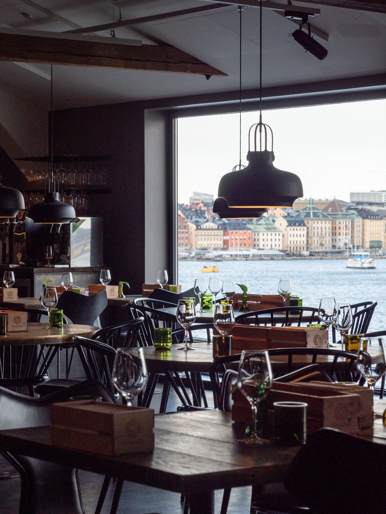Restaurant interior with set wooden tables and pendant lights, overlooking the water and Stockholm through a large window.
