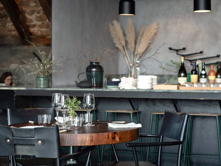 Restauranger i Stockholm. Spritmuseums restaurang, interiör. Ett bord står dukat för middag. Rustik inredning med varma färger; mörkt trä, gröngrå bardisk, grå tapeter och svarta stolar.