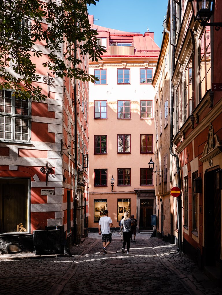 Narrow cobblestone alley in Gamla stan, Stockholm, lined with red and pink buildings as people walk through the sunlit street.