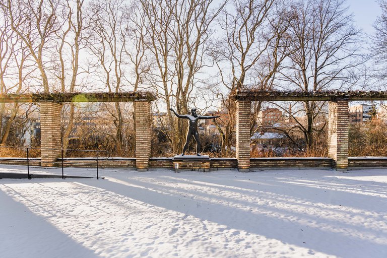 A bronze sculpture of Zeus or Poseidon standing in a snowy sculpture garden on a sunny day.