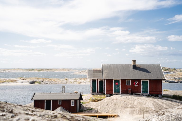 Red wooden cottages with green doors on rocky shores in the outer Stockholm archipelago, surrounded by smooth granite cliffs and open water beneath a blue sky.