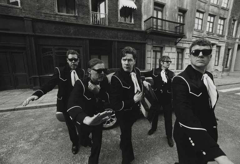 Five band members in matching black suits with white trim and ties, wearing sunglasses, striking dynamic poses on a city street; black-and-white photo.