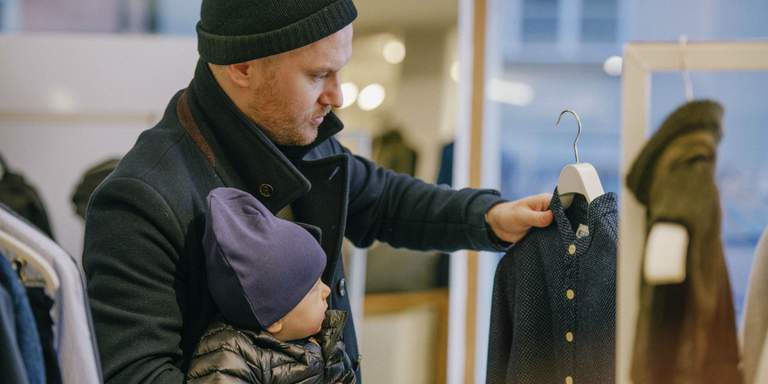 Shopping in Stockholm. A man browsing through clothes in a store. The man is wearing warm clothes and is carrying his child under one arm.