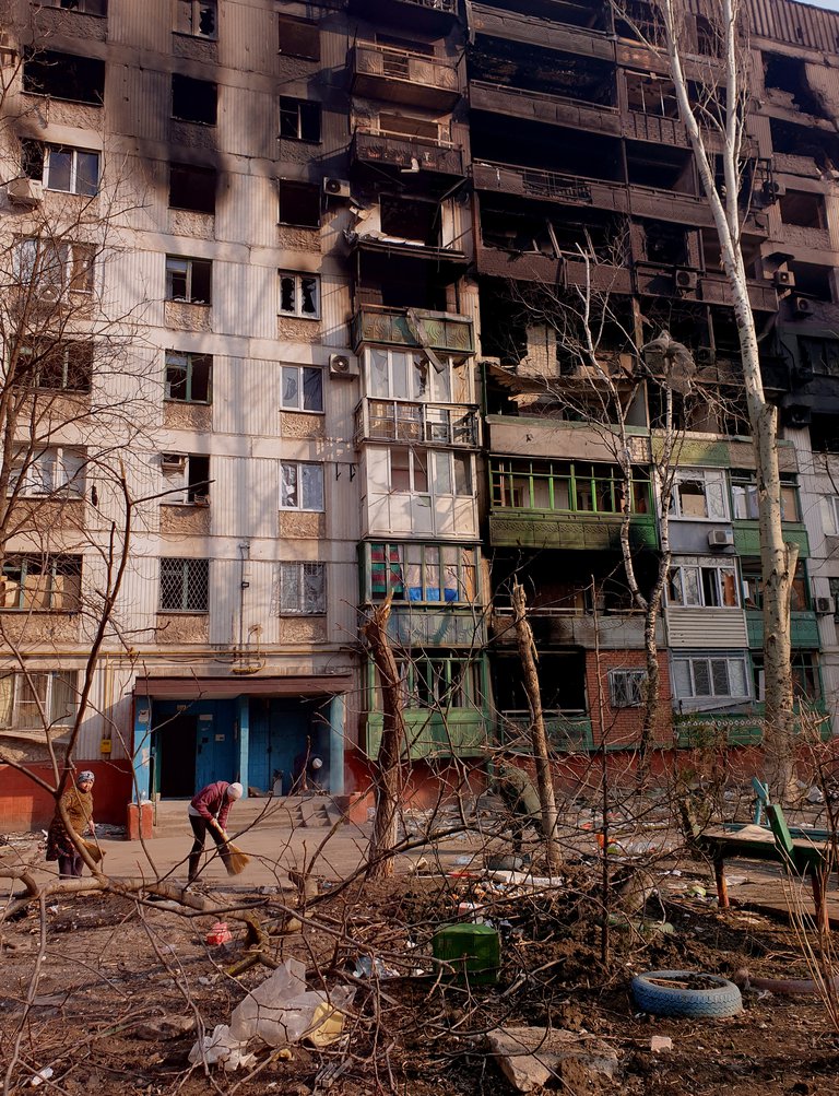 A damaged multistoried building. Two people cleaning in its courtyard.