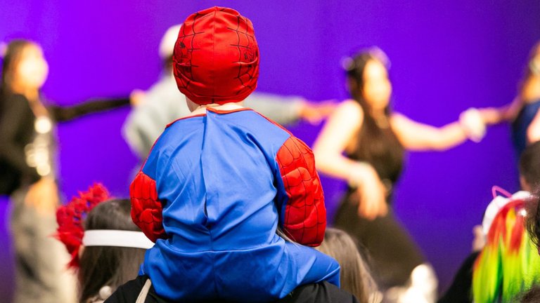 A child in a Spider-Man costume sits on an adult’s shoulders, watching a stage where several performers are blurred against a purple backdrop.