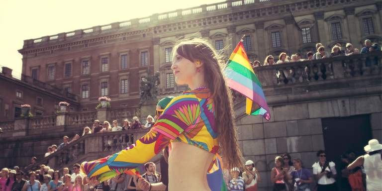 Summer in Stockholm, daytime. The annual Stockholm Pride-procession is walking by a crowd in front of Stockholm Palace. A woman in colorful clothes in carrying a rainbow flag.