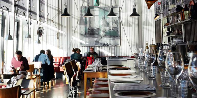 Parts of real boats and canoes are hanging high up in the ceiling, huge windows are letting in daylight in the restaurant. People are sitting at a few of the tables and a modest bar is visible in the foreground.