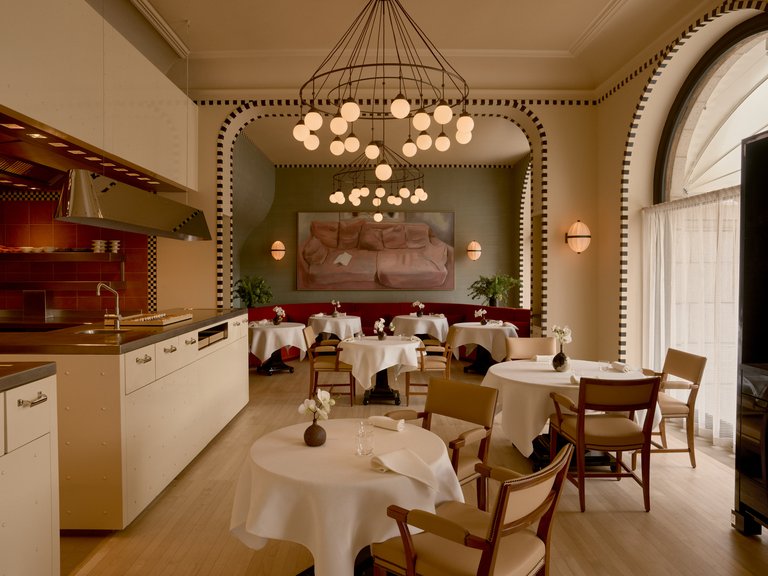Elegant restaurant interior with white tablecloths, an open kitchen, tall arched windows, and decorative chandeliers.