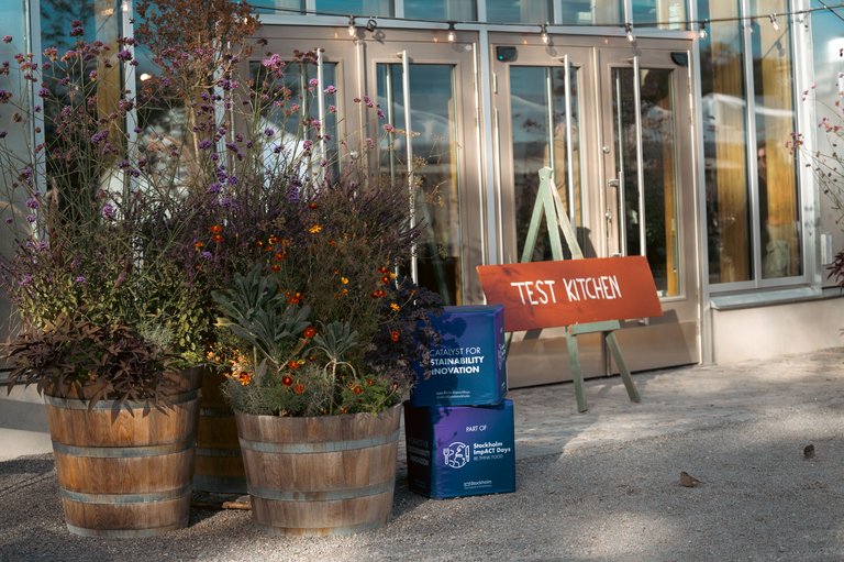 Entrance with wooden barrel planters and a “TEST KITCHEN” sign; two Stockholm Impact Days boxes by the door.