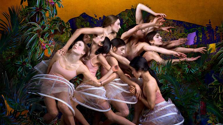 A group of young women in undergarments in what looks like a jungle. They stand packed together while looking and reaching in different directions like a renaissance painting.