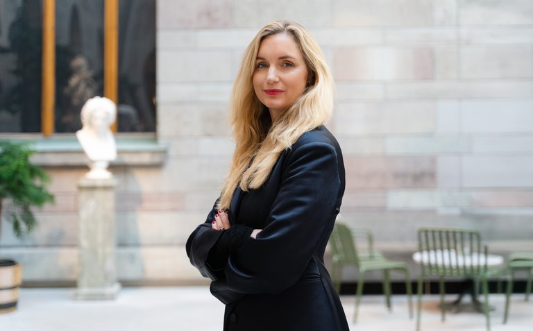 Portrait of Joanna Sundström standing in a museum, looking into the camera with a blurred background of exhibits.