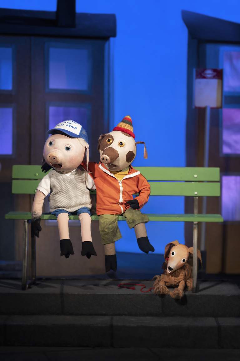 Three muppet dolls representing two pigs sitting on a bench and a dog near the bench.