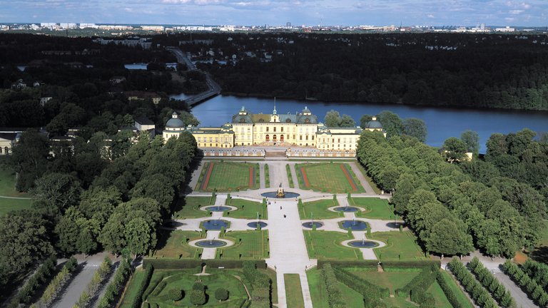 Areial shot of Drottningholm Palace in Stockholm, summer, daytime. A large white palace with a blossomiing garden in-front.
