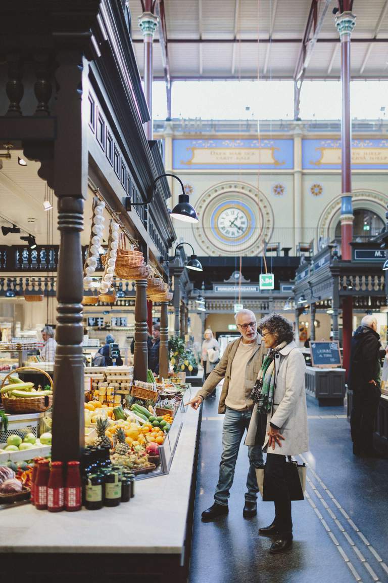 Östermalmshallen market hall in Stockholm. People are walking between the stalls, choosing what tou buy for dinner. The grand hall was bulit in the 1880s and has since then been a popular place to buy fresh vegetables, fish, cheese, cuts of meat and other delicacies.