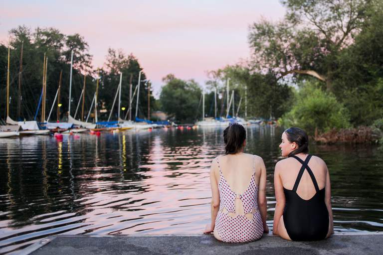 Two young women in swimming suits, backs to the camera, sit on the wharf at Långholmen after a swim in Lake Mälaren. There are several great places for a swim in Stockholm; even in the heart of the city!