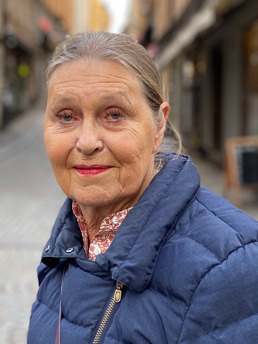 Meet the Locals. Ingegerd Wallén. En äldre kvinna, tittar in i kameran. Hon är klädd i en mörkblå jacka och bär på en vinröd axelväska