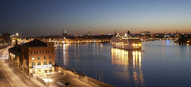 Färjor i Stockholm. Ett kryssningsfartyg ligger förtöjt utanför Slussen. Kväll. I bakgrunden syns Gamla stan. Ljuset från byggnaderna speglas i vattnet.