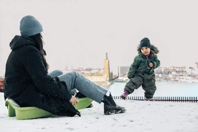 A toddler in a warm overall and knitted hat pulls their parent in a plastic sled. It's snowing and Stockholm City Hall is seen in the background.