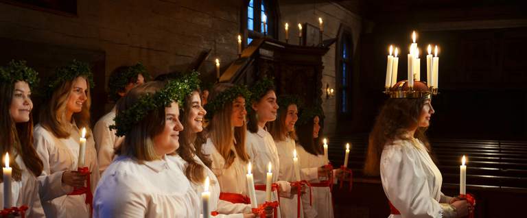 Saint Lucia performance with girls in white, traditional Lucia clothing and lit candles. Lucia has lit chandelier crown on her head.
