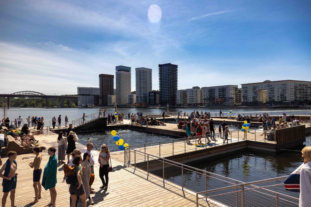 People at a sunny waterside bathing area with wooden piers; blue-yellow balloons, high-rises and a bridge in the background.