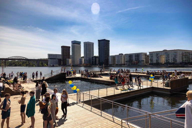 People at a sunny waterside bathing area with wooden piers; blue-yellow balloons, high-rises and a bridge in the background.