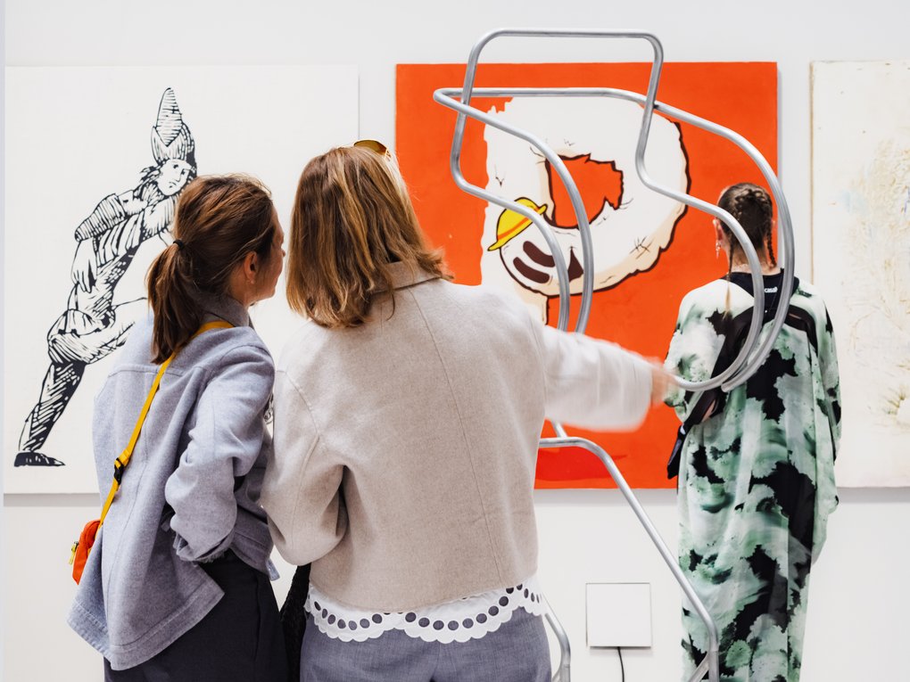 Visitors at an art fair viewing paintings and a sculptural metal form in a gallery space.