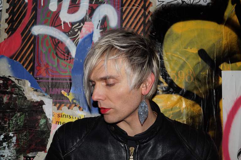 The artist Moto Boy with a graffiti-painted wall behind him.