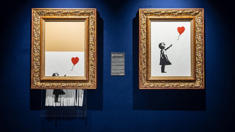 Banksy art hanging on a wall. One of the paintings is in the process of being shredded.