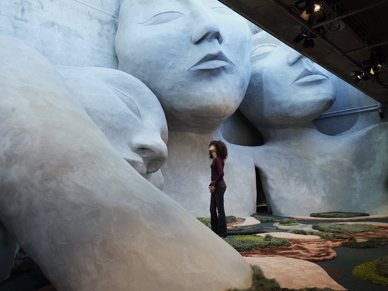 A person stands in an exhibition hall facing enormous sculptures of human faces and bodies made of light gray material. The monumental forms fill the space and create a calm, dreamlike at