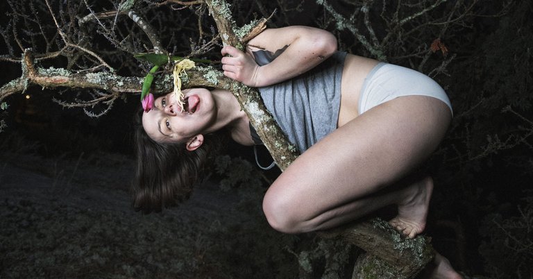 An actress wearing underwear climbing a tree branch. She's eating noodles lying directly on the branch.