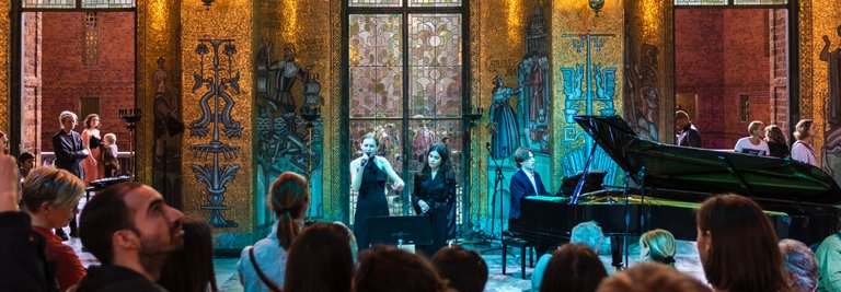 An audience stands gathered in the Golden Hall, which has large windows and golden walls, listening to a live performance. Two singers stand at microphones and a pianist plays the grand piano. The image conveys an atmospheric concert and cultural experience.