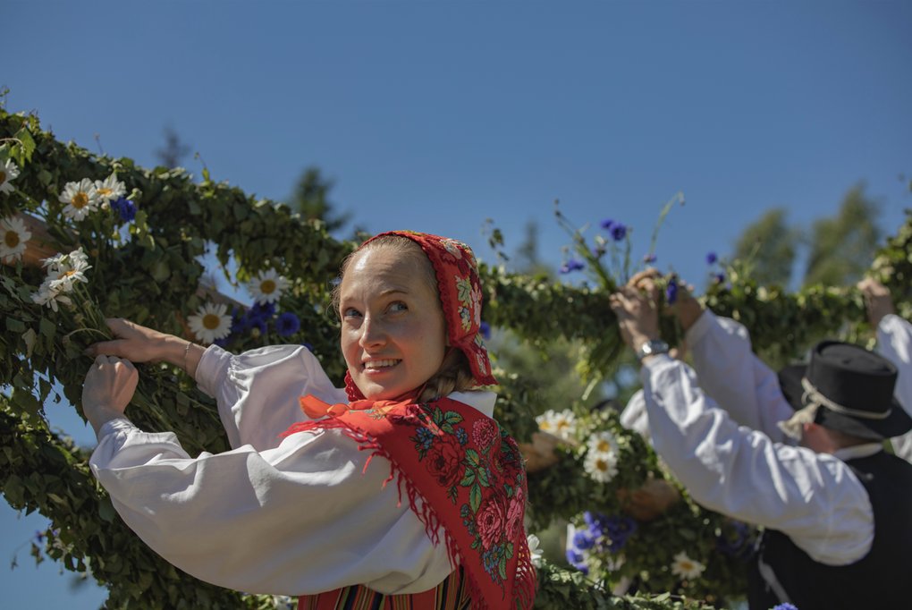 A woman in traditional folk costume decorates a maypole with greenery and flowers, with others helping in the background. She smiles toward the camera under a clear blue sky.
