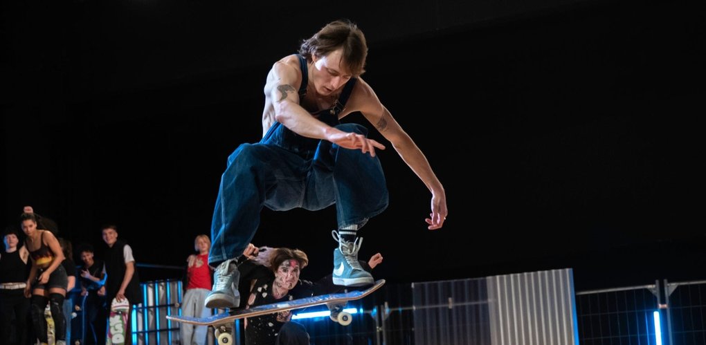 A skateboarder jumps high above an indoor ramp while other people watch in the background. The movement is dynamic, with bent knees and the board held close under the feet in mid-air. The image conveys speed, courage, and the energy of a skatepark setting.