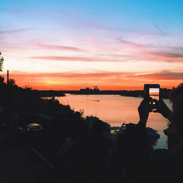 A person holding up a smart phone to take photos of an orange summer sunset in central Stockholm