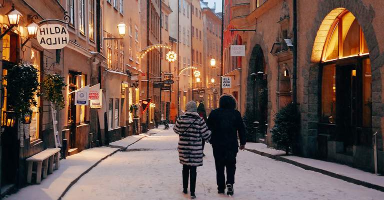 A couple walking on a snowy pedestrian street in Stockholm's old town in the evening. The street is narrow and lined with old, historical buildings in different shades of orange. Christmas lights shaped as stars are shining bright along the facades.