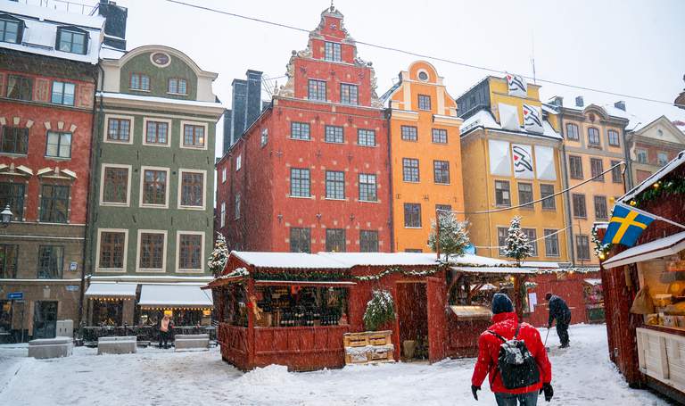 Red market stalls decorated with light strings, in a snowy square lined with colorful, old buildings