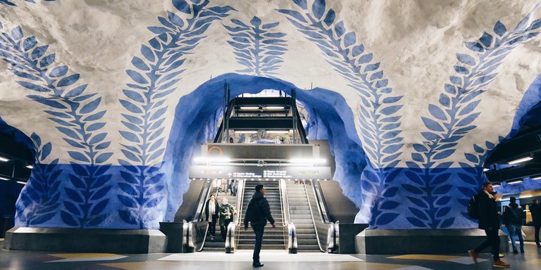 Blue and white leaf-pattern artwork at T-Centralen subway station in Stockholm, with escalators and commuters moving through the space.