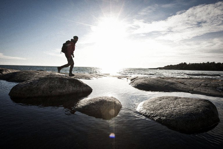 A hiker steps across smooth seaside rocks, backlit by the bright sun near the horizon.