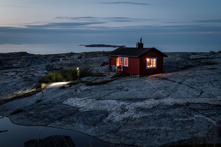 A cozy red cabin with lit windows sits on a rocky shoreline at twilight, a serene sea in the background.