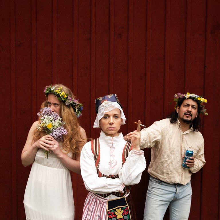 Three people in traditional Midsummer attire stand against a red wall.