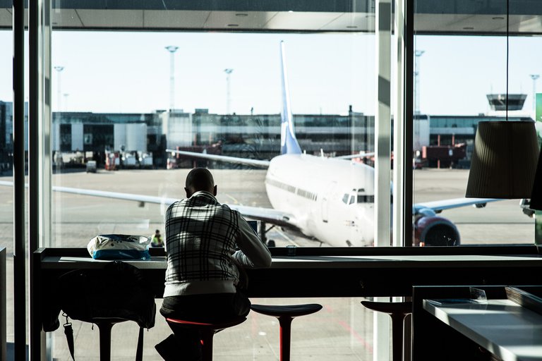 Reas till Stockholm. Arlanda flygplats. En man sitter vid ett fönster, med ryggen vänd mot kameran. Utanför syns landningsbanan på Arlanda och ett stillastående flygplan.