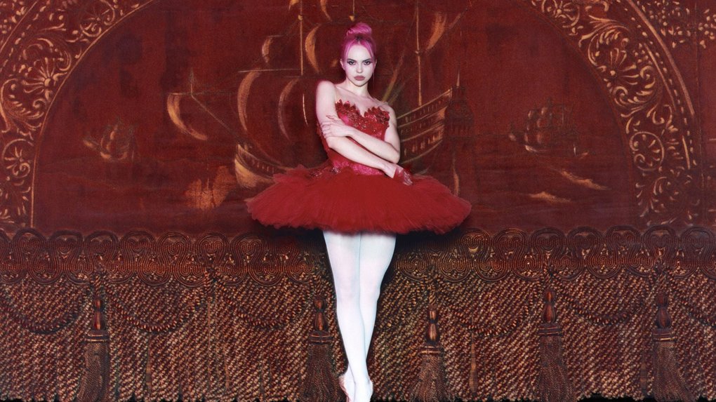 A person poses in a ballet outfit with a red tutu and white tights against an ornate golden-brown stage backdrop with patterns.