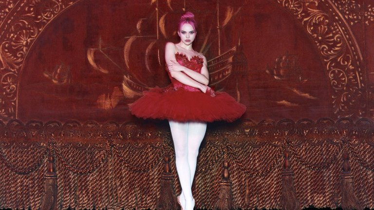 A person poses in a ballet outfit with a red tutu and white tights against an ornate golden-brown stage backdrop with patterns.