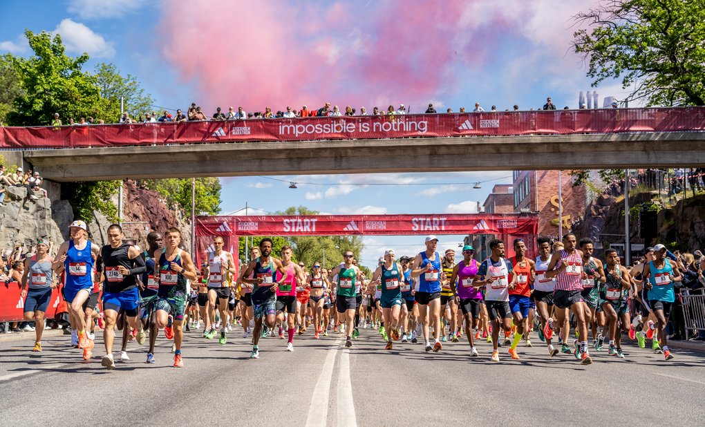 Runners start a marathon on a sunny day, passing under a bridge with a red banner that reads “Impossible is nothing.”