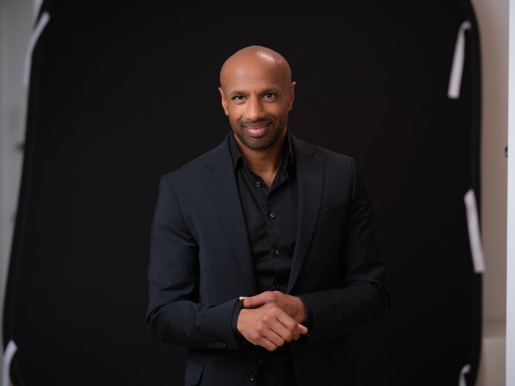 A man in a black shirt and suit jacket against a black backdrop.