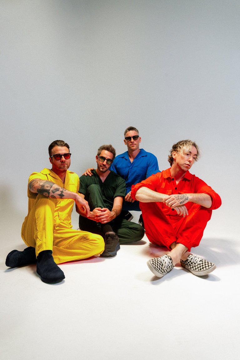 The members of All Time Low sit on the floor while dressed in colorful jumpsuits.