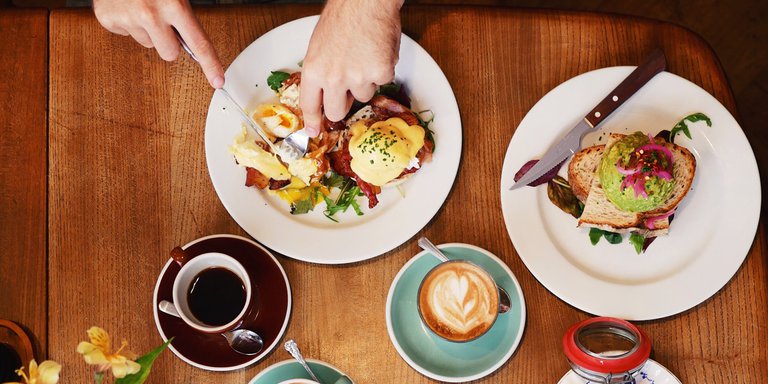: A bird´s eye view over a couple of people sharing brunch, a black coffee and a latte together with an Eggs Benedict and Avocado toast is on the table.