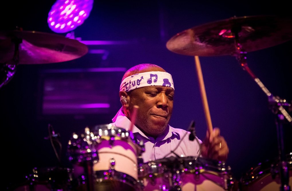 Drummer Billy Cobham plays intensely behind a drum kit on a stage lit with purple and pink lights. He wears a white headband with musical notes and holds drumsticks in motion. The image conveys energy, rhythm, and a live performance atmosphere.